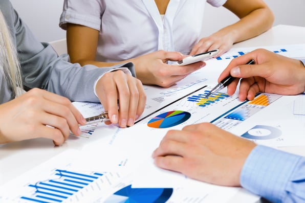 A close-up shot of several people's hands working together at a table. They're reviewing documents with charts and graphs, and one person is using a calculator.